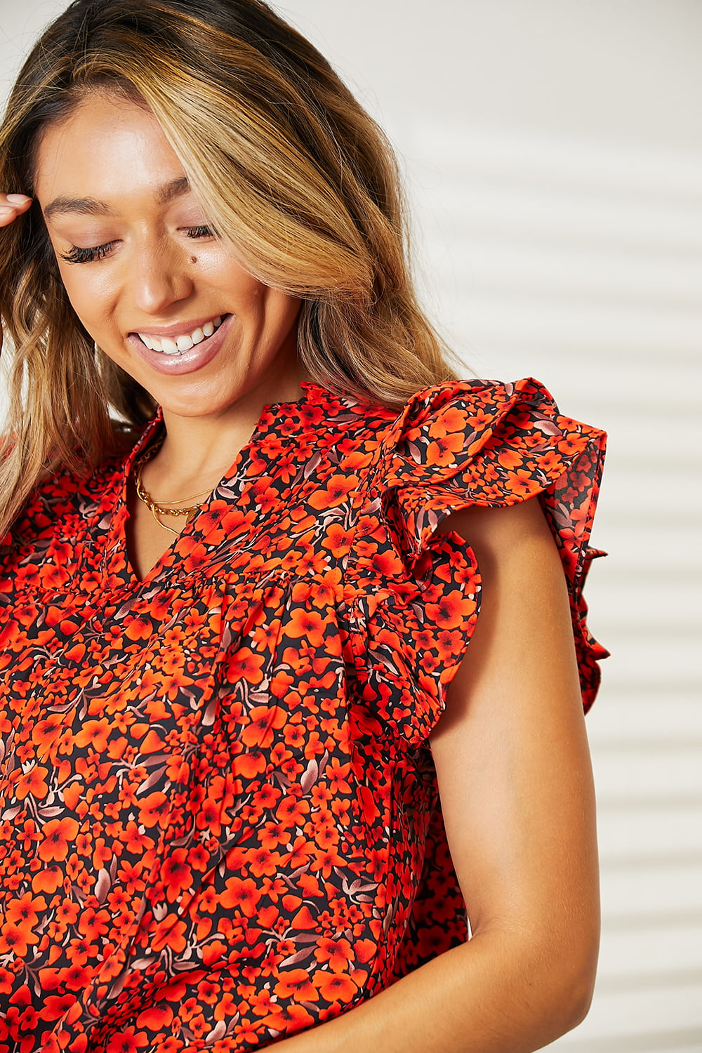 A woman wearing a chic Double Take Floral Flutter Sleeve Notched Neck Blouse and blue jeans stands smiling in a well-lit room with white walls and a curtain.