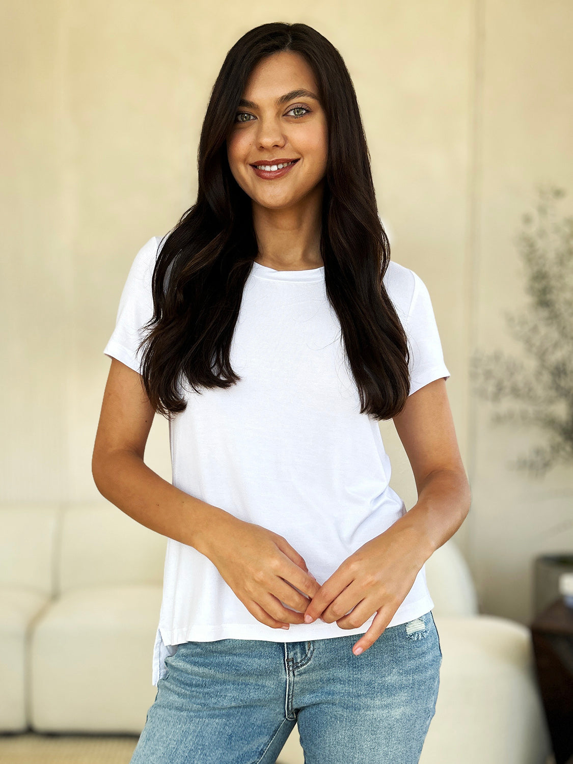 A woman stands next to an indoor plant, smiling, with one hand in her khaki pants pocket. In her pink Basic Bae Full Size Round Neck Short Sleeve T-Shirt and gold jewelry, she radiates casual elegance.