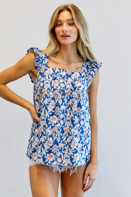 A woman in a vibrant summer wardrobe stands against a plain background, wearing the Floral Printed Ruffle Sleeveless Top paired with white shorts, with one hand casually tucked in her pocket.