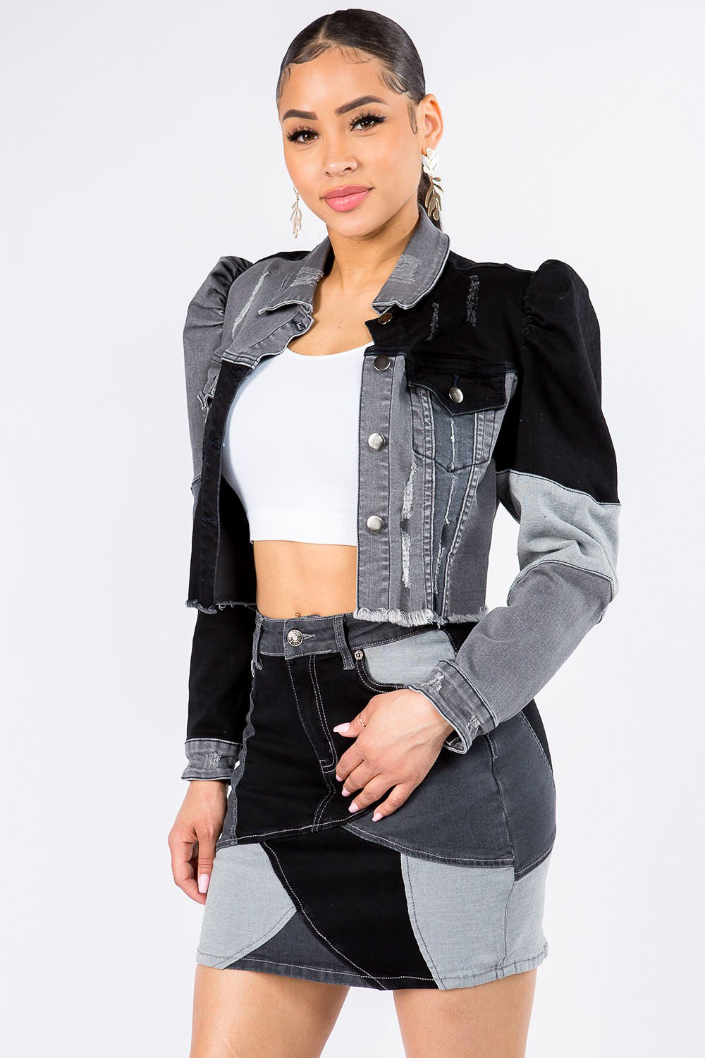 A woman is wearing the American Bazi Button Up Cropped Patchwork Denim Jacket, featuring puff shoulders and a raw hem, paired with a matching skirt in black, grey, and white. Underneath, she sports a white crop top while standing against a plain white background.