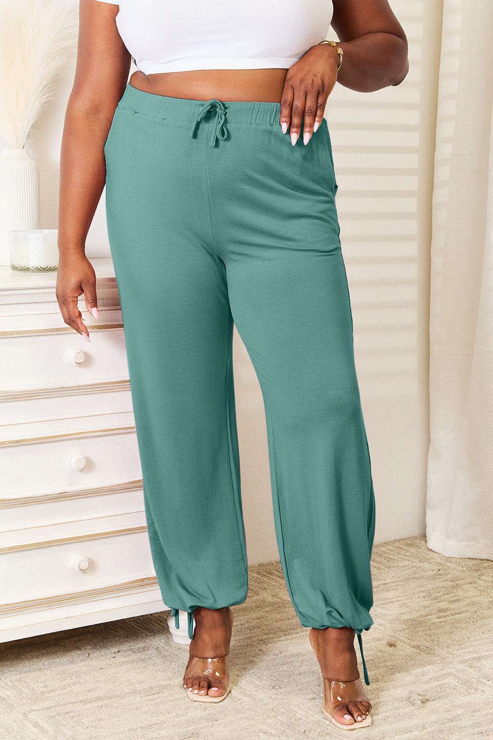 Person wearing Basic Bae Full Size Soft Rayon Drawstring Waist Pants with Pockets in teal, standing next to a white dresser with hands in pockets. These comfortable and stylish pants represent versatile everyday wear.