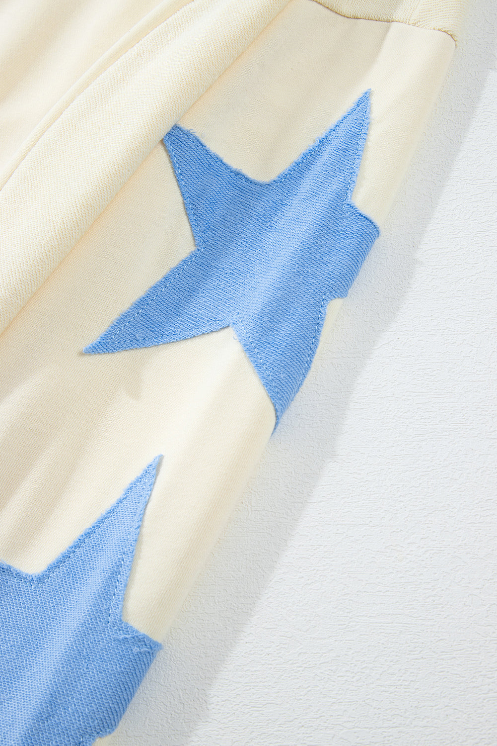 A person with long, wavy, light brown hair stands facing away, wearing the Beige Star Patchwork Exposed Seam Oversized Sweatshirt paired with denim shorts. The background is a plain, light peach wall, adding an edgy vibe to the look.