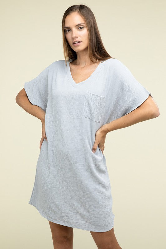 A woman wearing a loose light brown Woven Airflow V Neck T-Shirt Dress with Pockets stands against a plain background. Made from breathable fabric, this summer dress includes pockets where her hands rest as she smiles slightly.