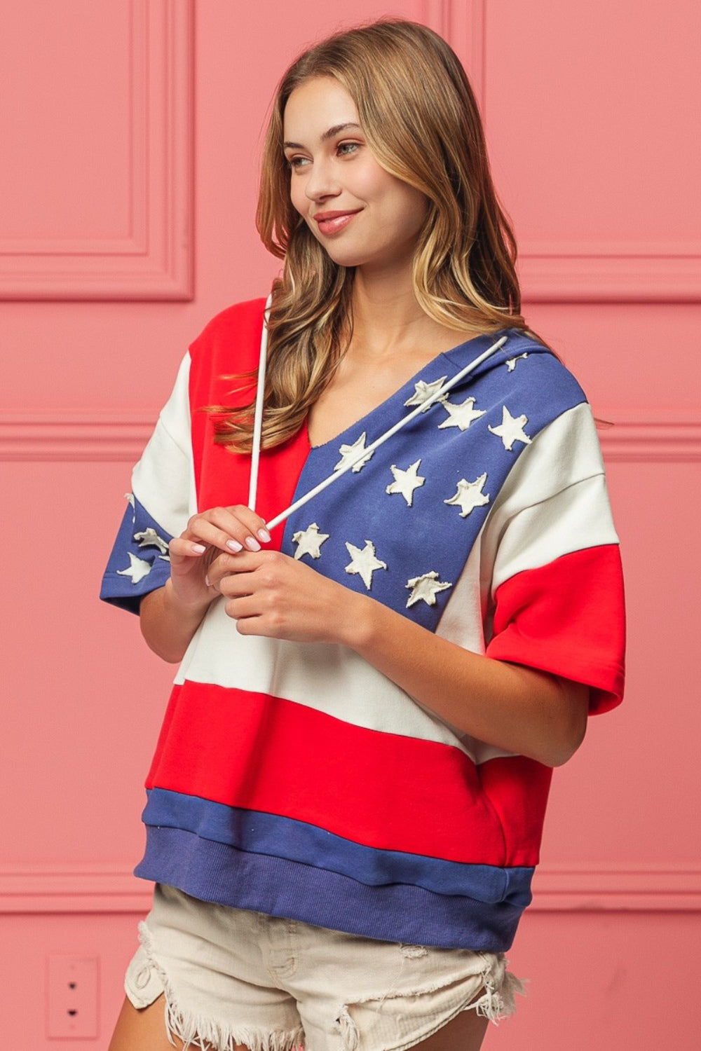 Woman wearing a BiBi American Flag Theme Hoodie, standing against a pink background.