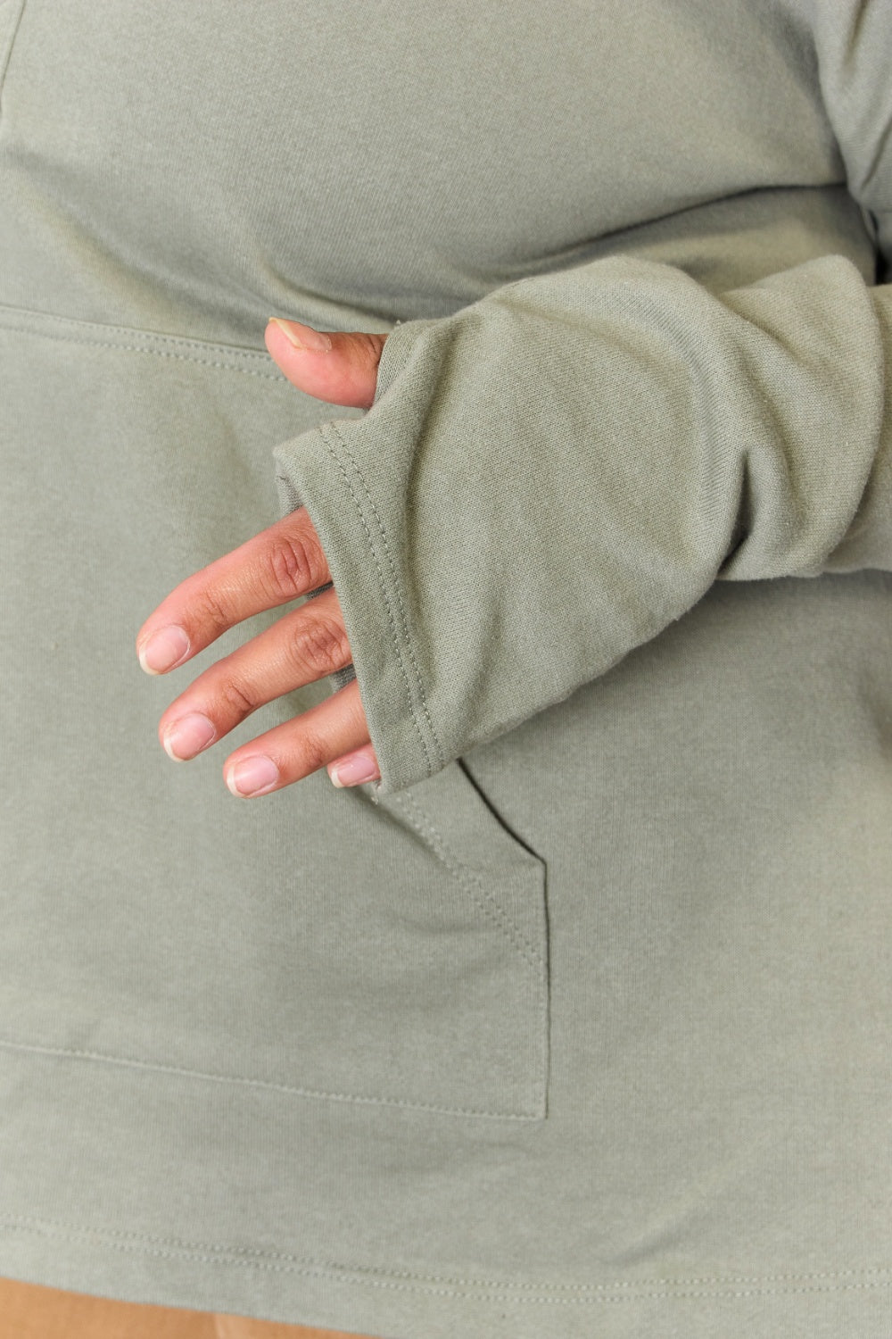 A person stands with hands in the pockets of their light green Culture Code Full Size Half Button Hoodie and brown pants. A plant is visible on the left side of the image.