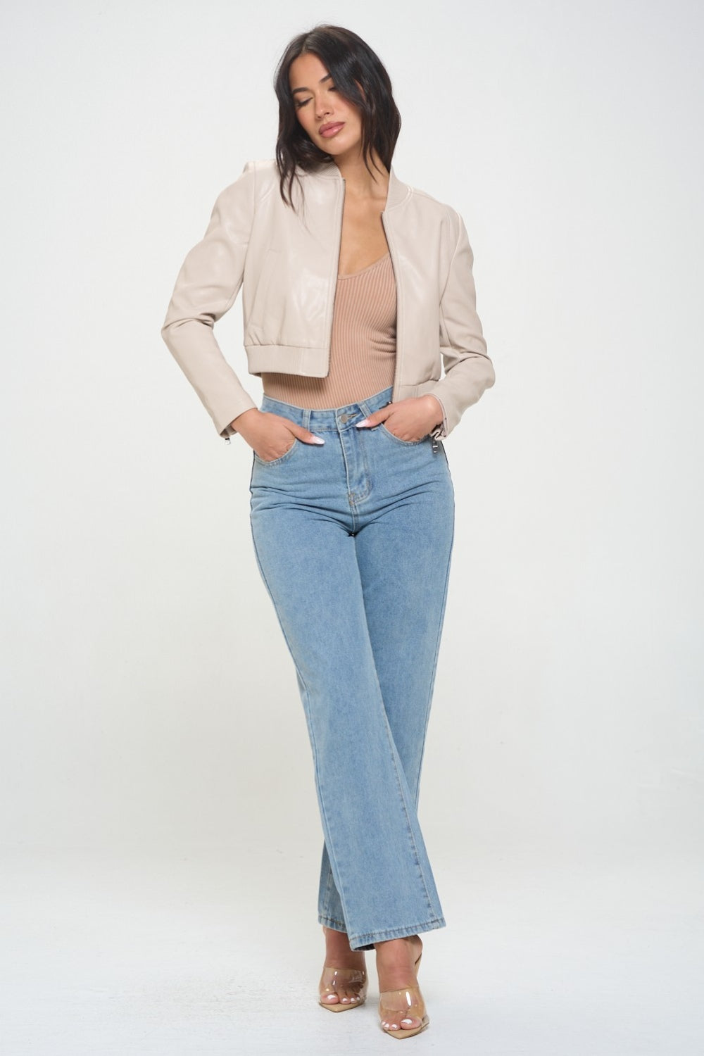 A woman wearing a Coalition LA Zip Up Cropped Bomber Jacket over a ribbed beige top and light blue jeans stands facing the camera, embodying sustainable fashion.