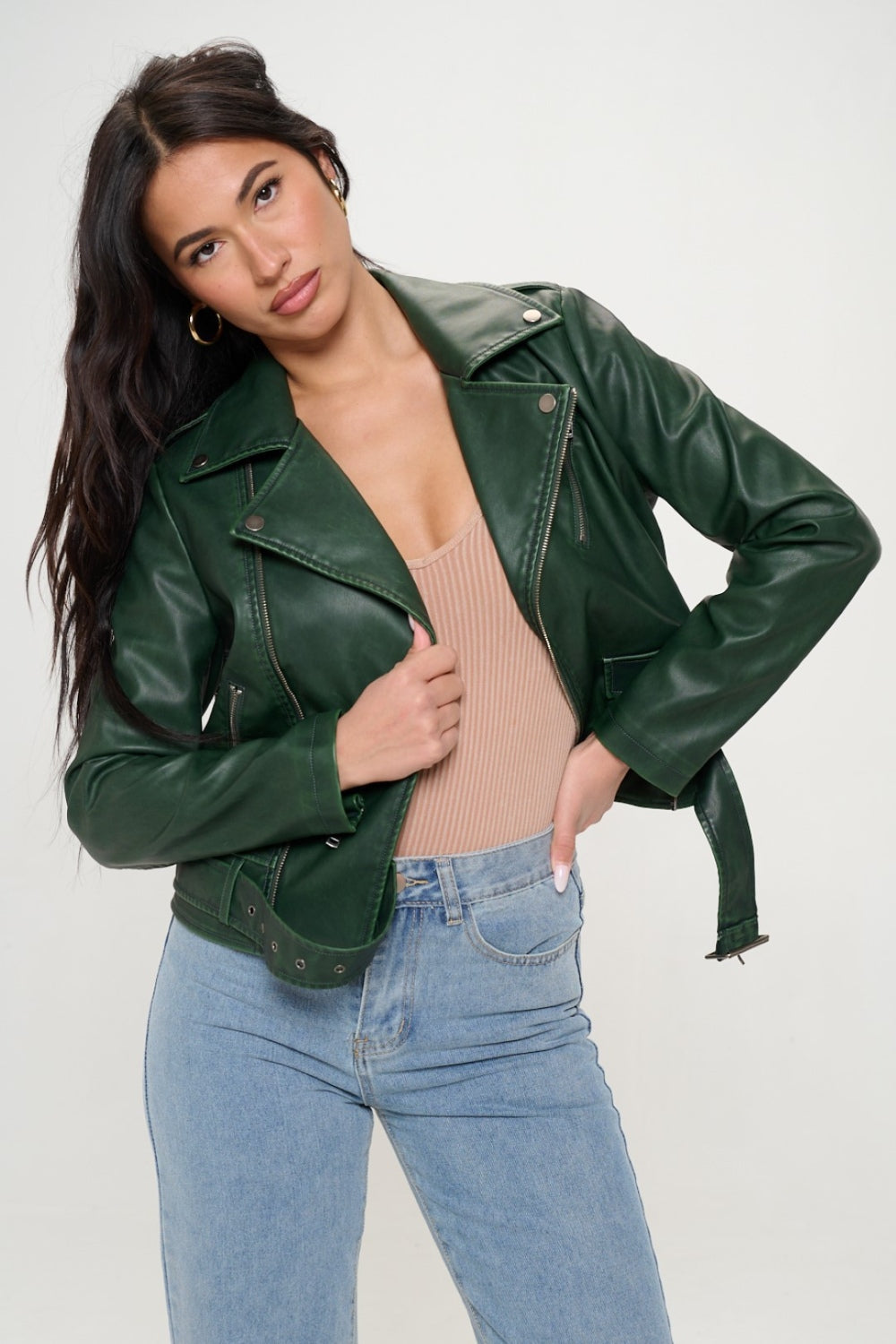 A person with long dark hair wearing a green Coalition LA Zip Up Biker Jacket with Belt and blue jeans stands against a plain background.