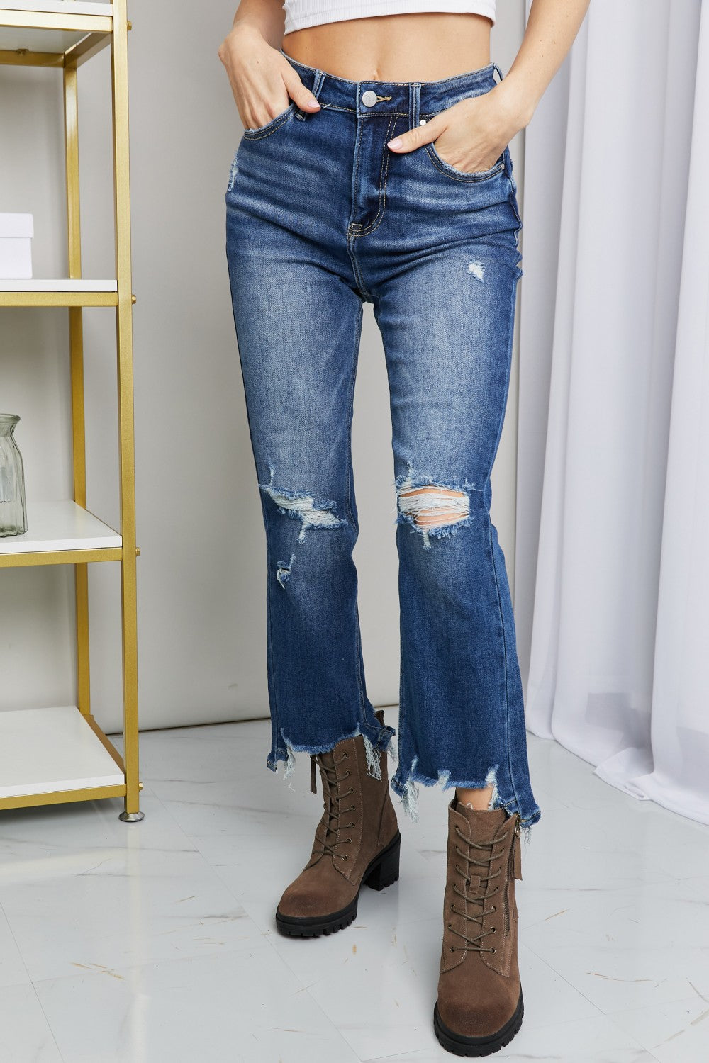 A person wearing RISEN Frayed Hem Distressed Cropped Jeans and brown ankle boots with a block heel stands in front of a white curtain next to a gold shelving unit.