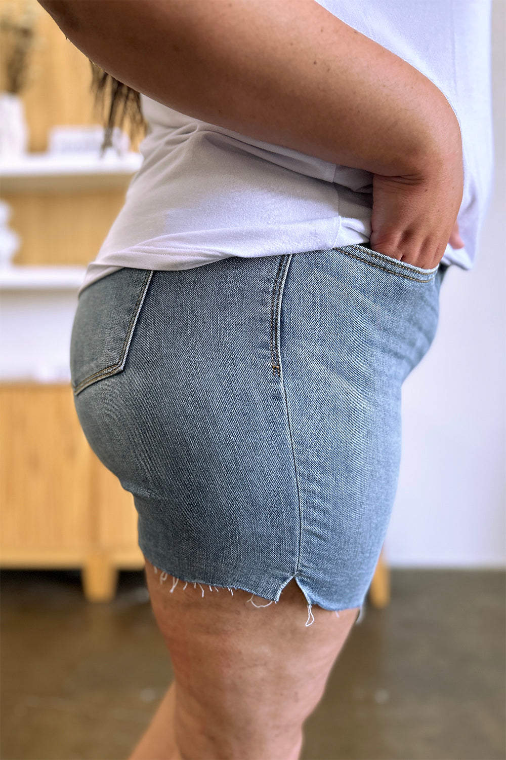 Side view of a person wearing a white shirt and Judy Blue Full Size High Waist Raw Hem Denim Shorts, standing indoors near a wooden cabinet. Summer fashion vibes radiate through the casual ensemble.