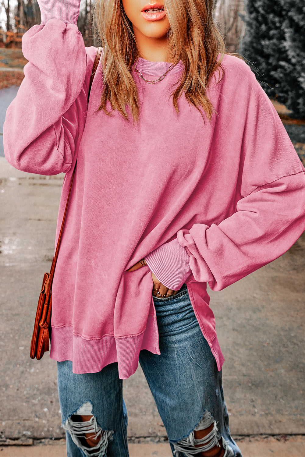 A person with wavy blonde hair, wearing a Pink Drop Shoulder Ribbed Trim Oversized Sweatshirt, blue jeans, and holding a brown bag stands outdoors with their back to the camera.