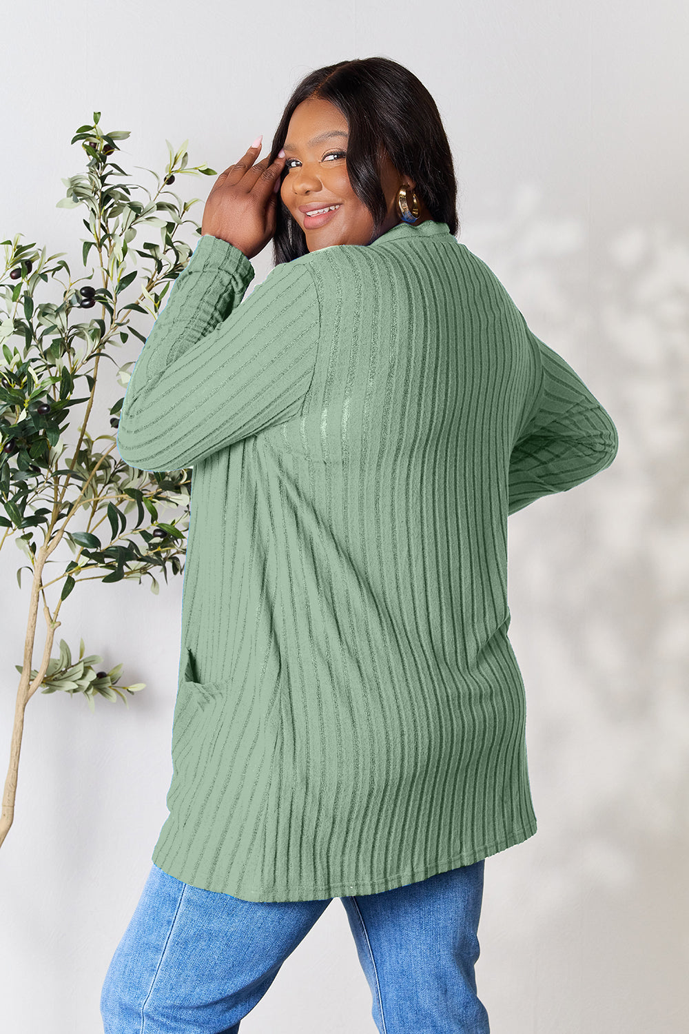 A woman in a Basic Bae Full Size Ribbed Open Front Cardigan with Pockets and jeans, made from stretchy material, stands in front of a plant, smiling slightly and looking to the side.