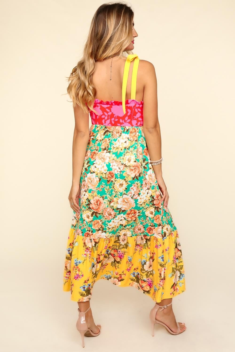 A person wearing the Haptics Floral Color Block Maxi Dress with Pockets, featuring a vibrant floral print and a color block maxi skirt in pink and yellow hues, stands smiling against a beige background.