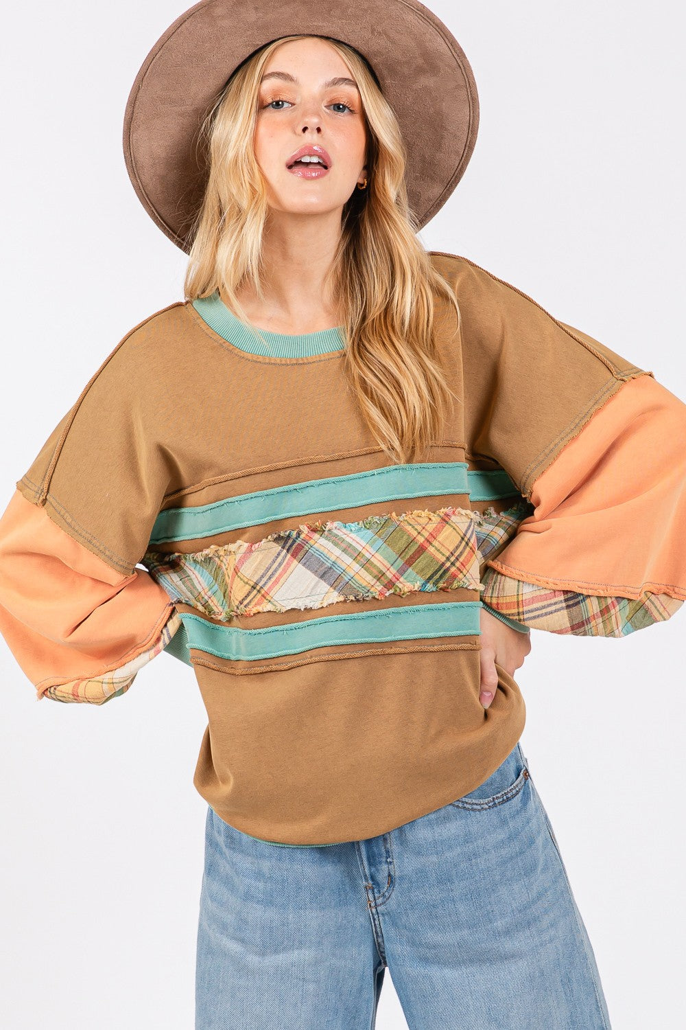 A person wearing a brown hat and the SAGE + FIG Mineral Wash Raw Edge Color Block Sweatshirt, featuring a patchwork design with plaid print contrast and solid color panels, smiles while posing with one hand in their pocket.