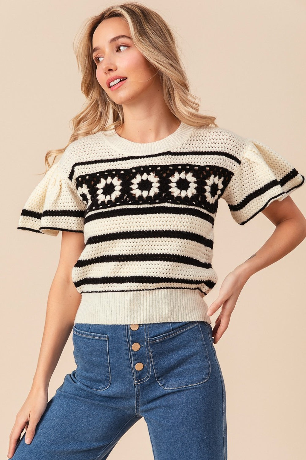 A woman sporting the BiBi Granny Square Short Sleeve Striped Sweater, which features cream and black stripes with floral crochet patterns on the chest, paired with blue jeans. She has light hair and is posing against a neutral background.