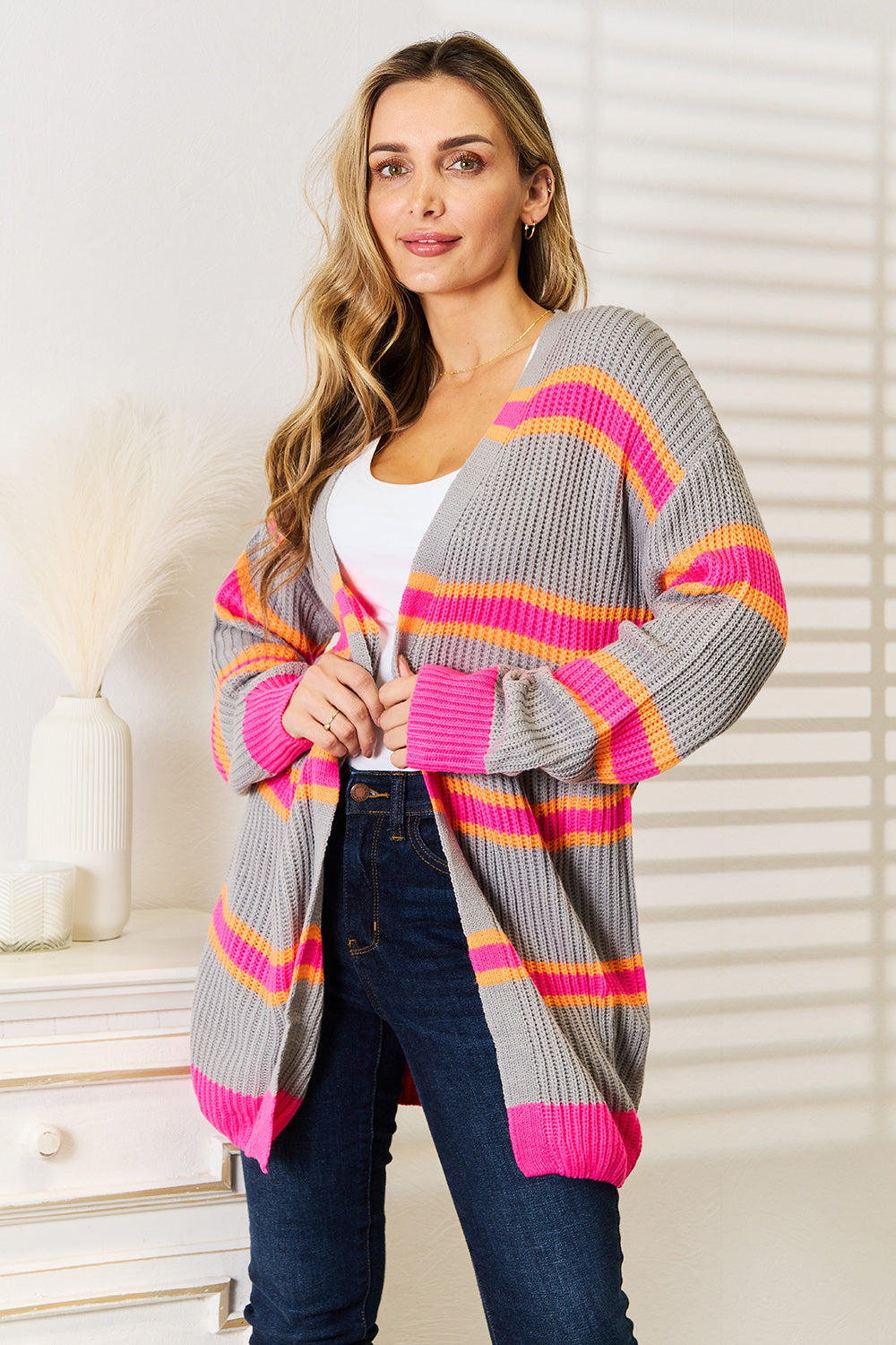 A woman stands against a white wall, smiling with one hand on her hip. She wears the Woven Right Ribbed Long Sleeve Cardigan, featuring pink and orange stripes, over a white top and dark jeans—an excellent choice for versatile layering.