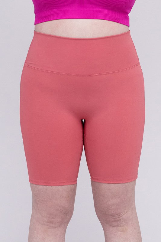 Against a plain background, a person showcases the Plus Athletic High Rise Biker Shorts in coral, crafted from high-quality athletic fabric and paired with a pink top.