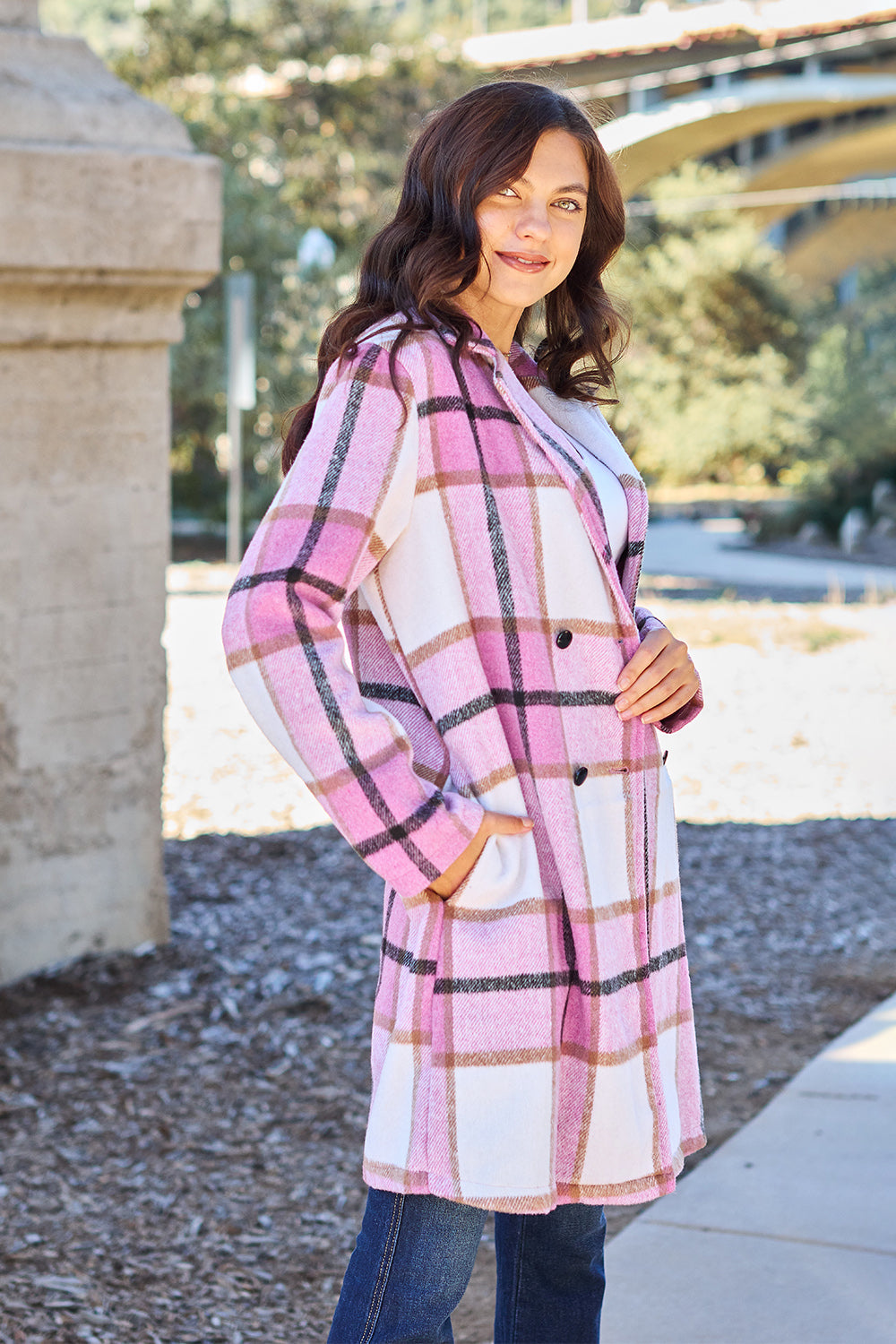 A woman stands on a sidewalk wearing the Double Take Full Size Plaid Button Up Lapel Collar Coat in pink and white over a white top and blue jeans, with her right hand lightly touching her hair. The polyester fabric of the coat makes it durable and perfect for an easy machine wash cold.