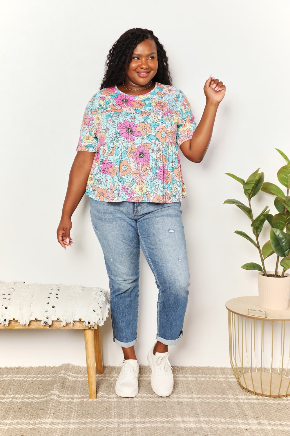 A person wearing a Double Take Floral Round Neck Babydoll Top and jeans stands with one hand in their pocket, with a plant in the background.