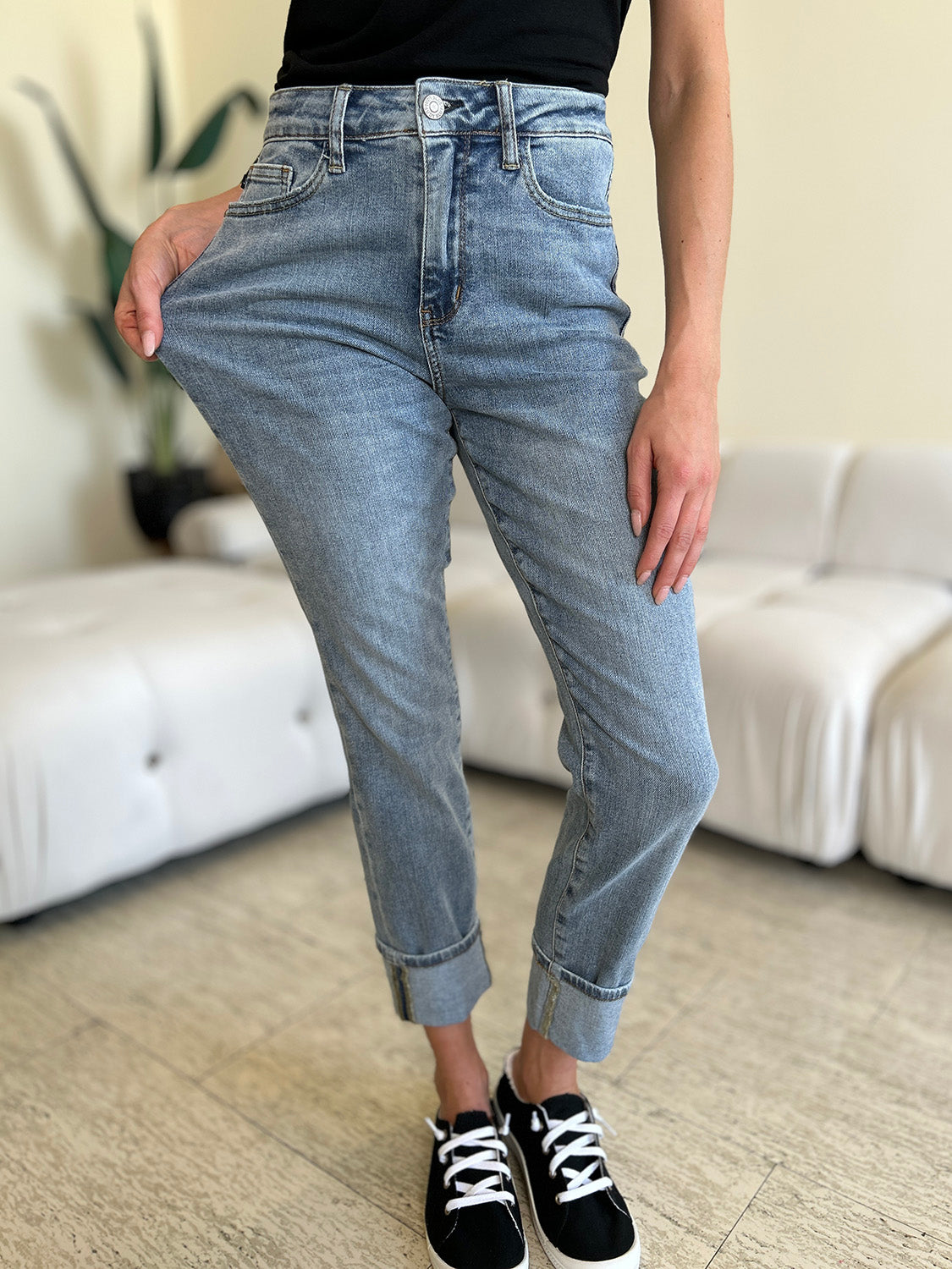 A person wearing Judy Blue Full Size High Waist Cuff Hem Jeans and white slip-on shoes stands in a living room with a white couch and plants in the background.