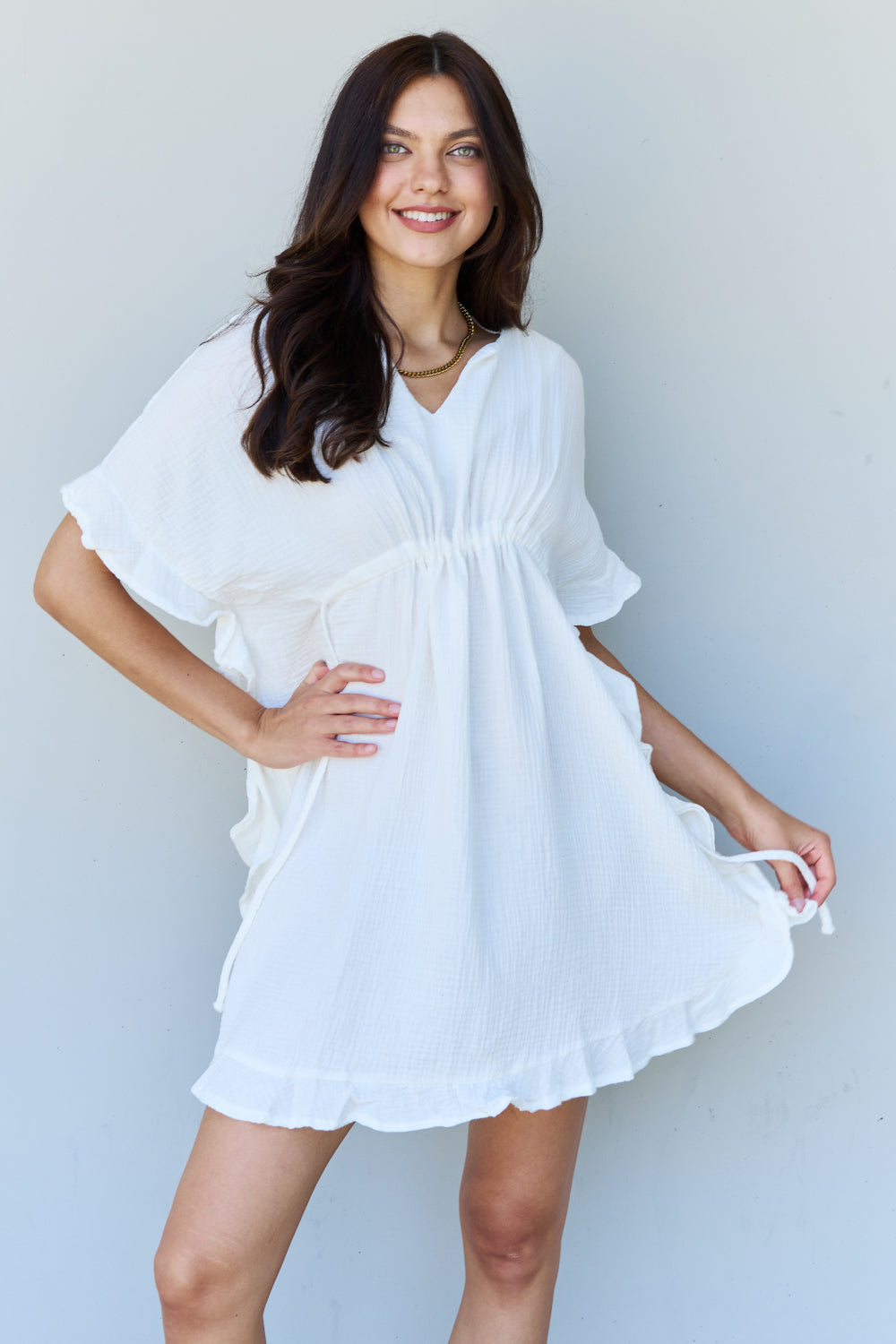 A person with long dark hair smiles while posing in the Ninexis Out Of Time Full Size Ruffle Hem Dress with Drawstring Waistband in White against a plain background.