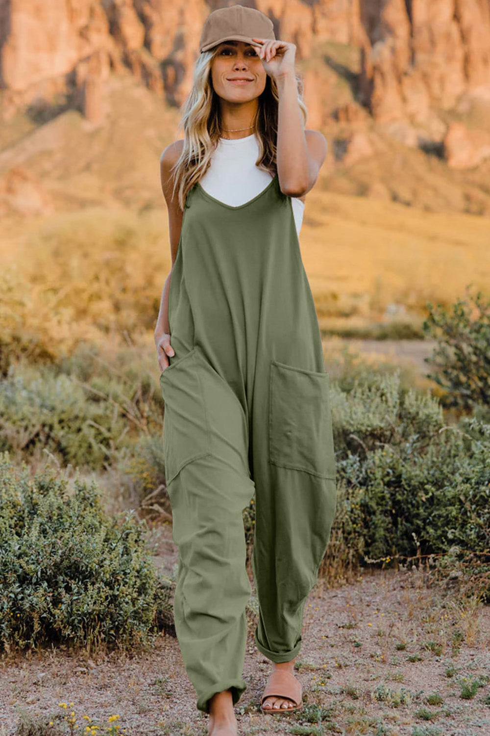 Person wearing a Double Take Full Size V-Neck Sleeveless Jumpsuit with Pockets and sandals, walking outdoors on a dirt trail with bushes in the background.