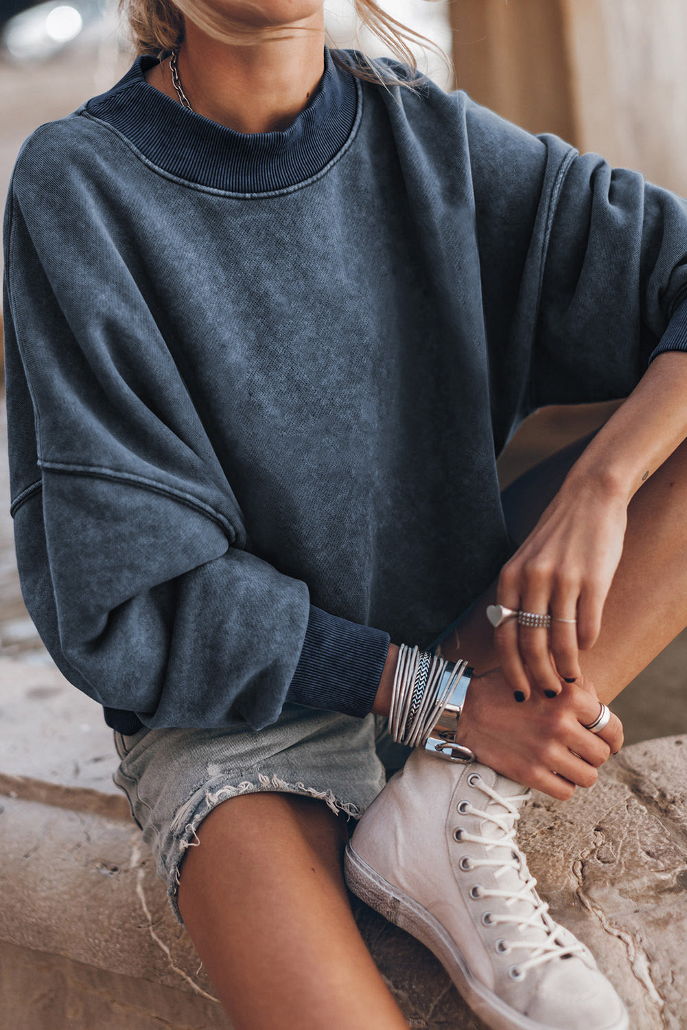 A person with long blonde hair is wearing a sky blue drop shoulder crew neck pullover sweatshirt and light denim shorts, standing outdoors with their back to the camera.