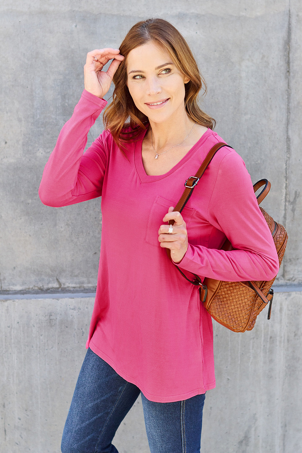 A woman with light brown hair stands against a concrete wall, wearing the Basic Bae Full Size V-Neck Long Sleeve Top in olive green and blue jeans, with a white backpack on her shoulder. Her outfit exudes basic style and is made from polyester, ensuring it’s both comfortable and suitable for machine wash cold. She is looking at the camera.