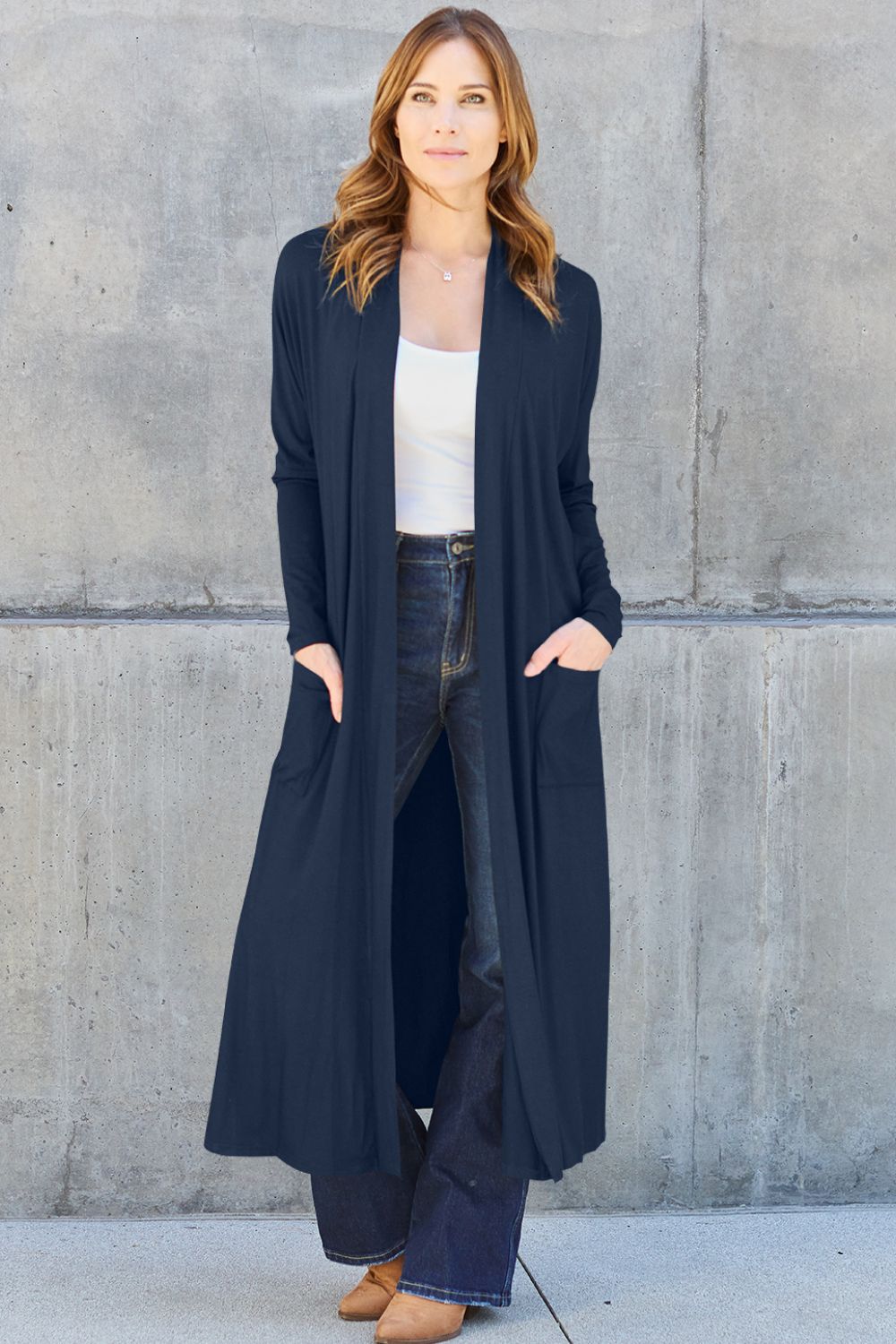 A woman stands against a concrete wall, wearing the Basic Bae Full Size Open Front Long Sleeve Cover Up made from rayon spandex, a white top, blue jeans in her perfect size, and brown shoes with her hands in her pockets. Machine wash cold for best results.