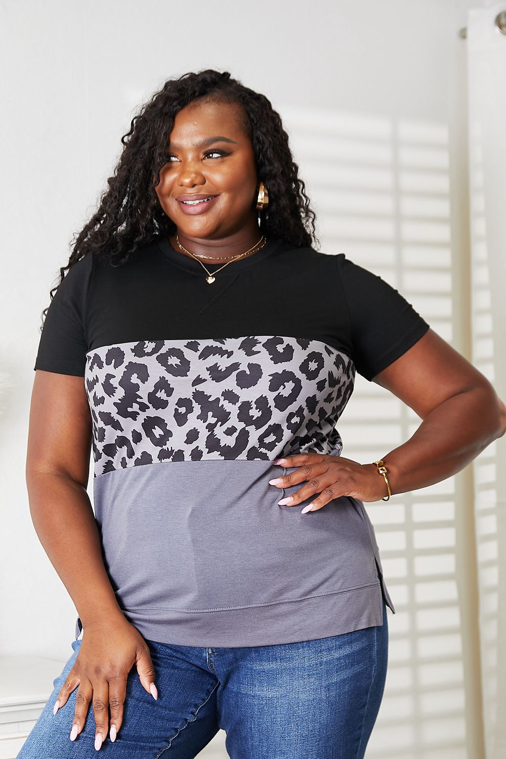 A person wearing a Double Take Leopard Print Color Block Short Sleeve T-Shirt and blue jeans stands indoors, smiling.