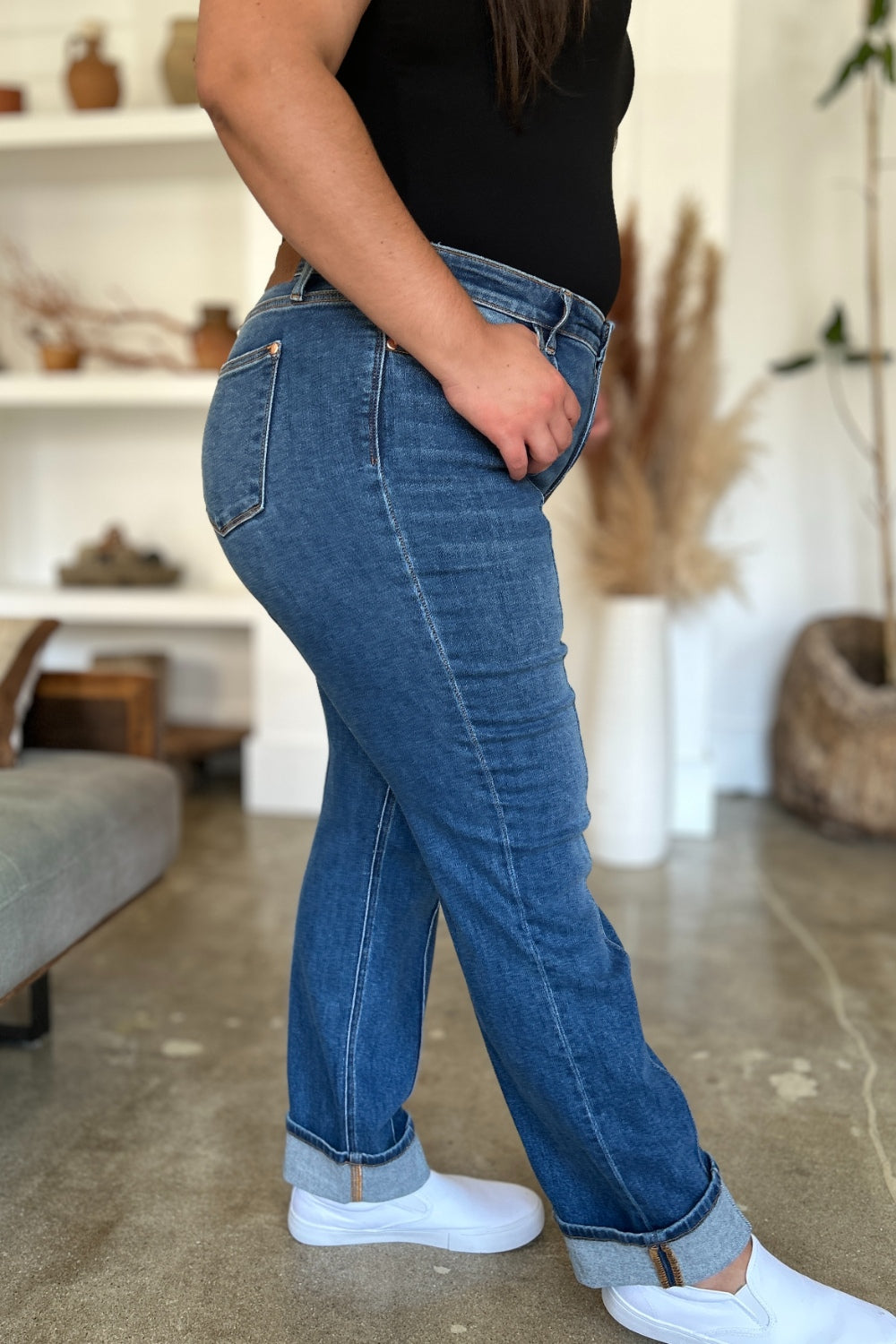 Two women stand side by side indoors, both wearing black tank tops, Judy Blue Full Size High Waist Front Seam Detail Straight Jeans, and white shoes. They are smiling and posing for the camera. A plant and shelves with decorations are in the background.