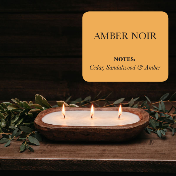 A luxurious 3-wick candle holder made from wood showcases three radiant candles surrounded by verdant foliage. The inscription reads "Amber Noir: Notes - Cedar, Sandalwood & Amber," embodying the elegance of our 3-Wick Dough Bowl Soy Candle - Amber Noir collection.