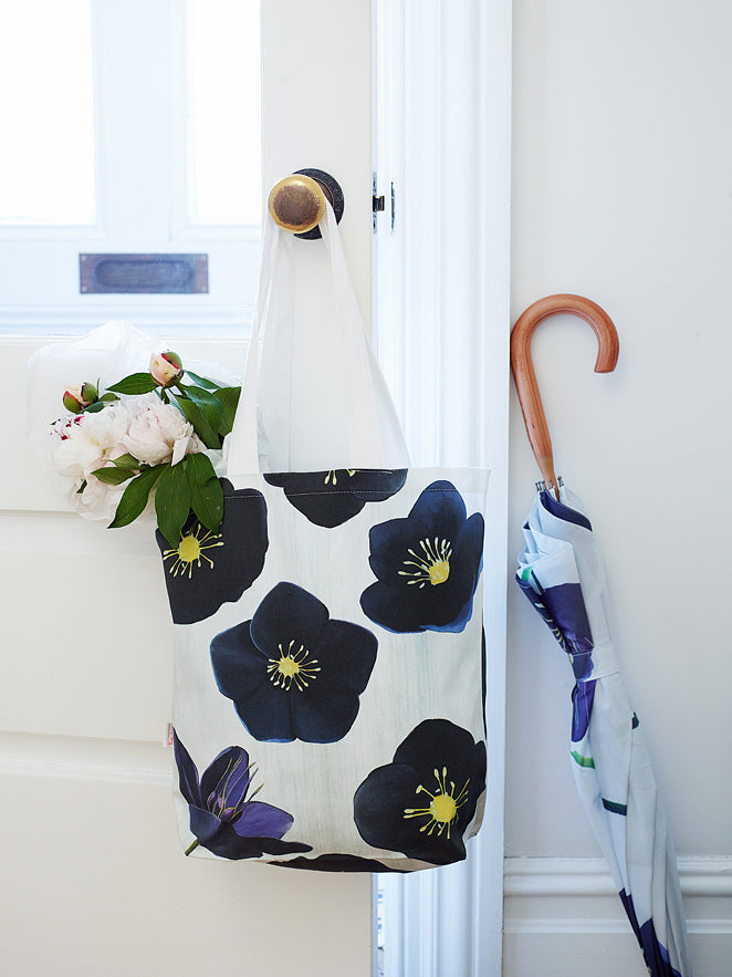 The Tote Bag: Black Hellebore on Snow, a chic cotton canvas piece with an elegant floral design, hangs on a doorknob beside a bouquet of Black Hellebore flowers. An umbrella featuring the same floral pattern leans gracefully against the wall.