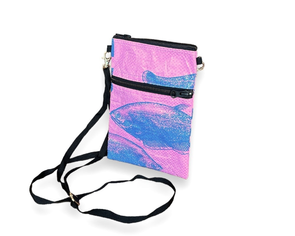 The Breezy Crossbody showcases a vibrant blue fish print on a pink background, crafted from upcycled materials. It includes two black zippered compartments and an adjustable black strap.