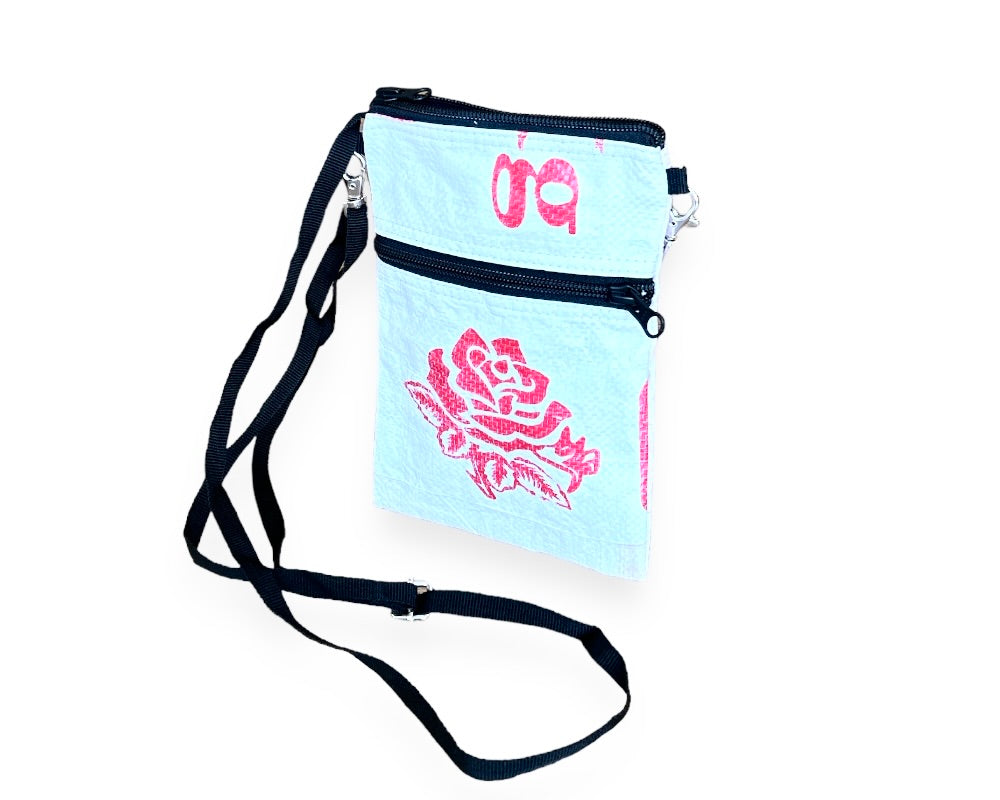 The Breezy Crossbody showcases a vibrant blue fish print on a pink background, crafted from upcycled materials. It includes two black zippered compartments and an adjustable black strap.