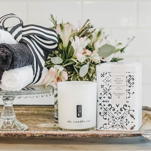 The Belgian Linen - Fresh Linen Soy Candle, along with a decorative tin, folded towels, and a flower arrangement, is elegantly displayed on a silver tray, exuding the soothing scent of fresh linen.