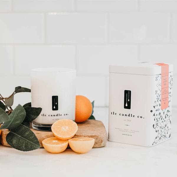 A "Citrus Tree - Orange Citrus Soy Candle" sits on a countertop next to a decorative box, with vibrant orange citrus fruits and lush green leaves nearby, against a backdrop of white tiled walls.