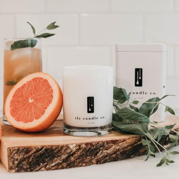 The Grapefruit Mojito - Grapefruit and Mint Soy Candle is showcased on a white surface alongside a tin of tea, fresh mint, grapefruit slices, and a candle wick trimmer. The soy candle fills the air with its essential oil fragrance, as matches lie in readiness to ignite the tranquil setting.