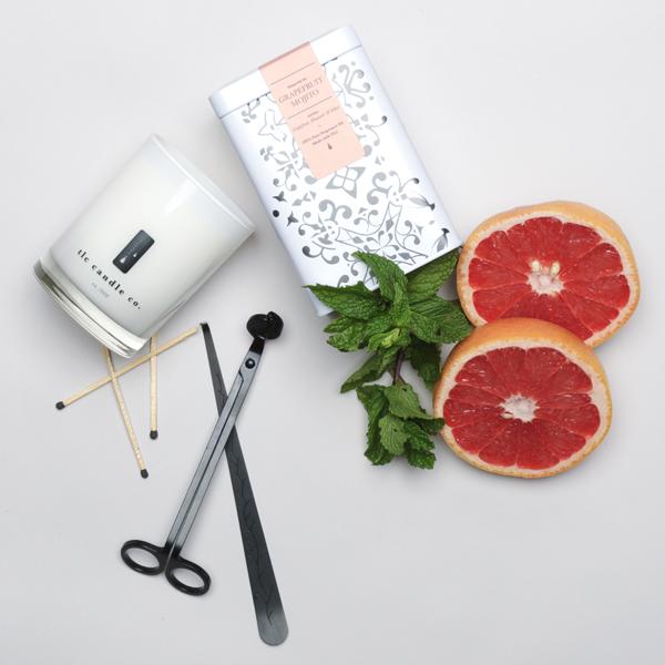 The Grapefruit Mojito - Grapefruit and Mint Soy Candle is showcased on a white surface alongside a tin of tea, fresh mint, grapefruit slices, and a candle wick trimmer. The soy candle fills the air with its essential oil fragrance, as matches lie in readiness to ignite the tranquil setting.