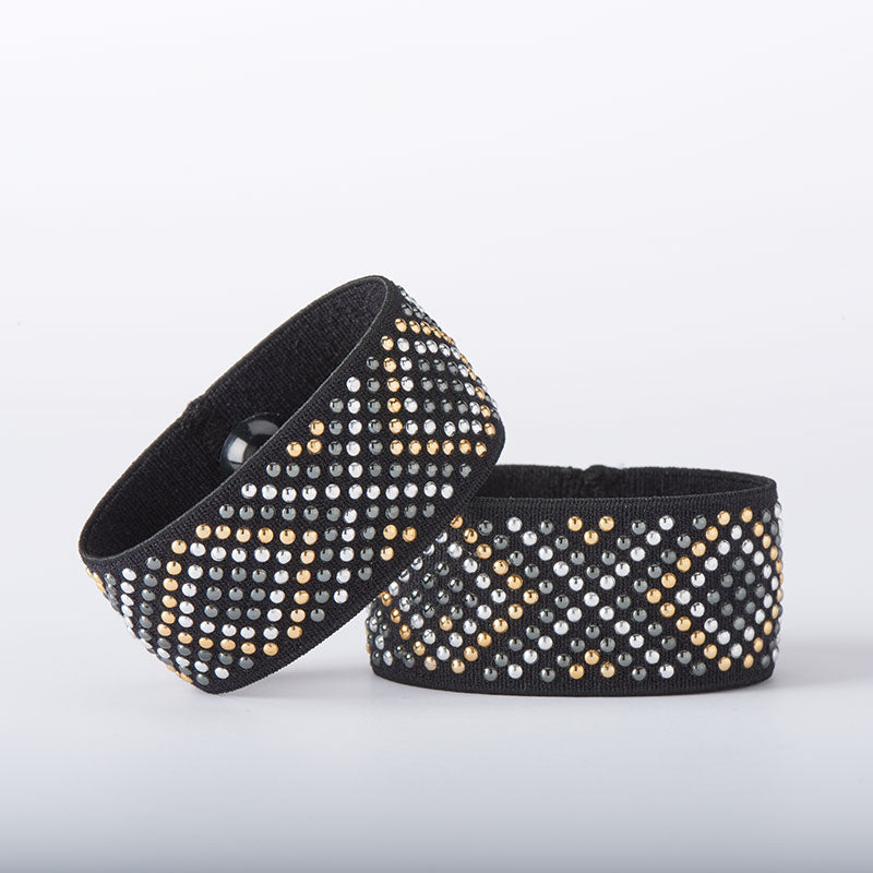 The Cecilia Duo Nausea Relief Bracelets, featuring two black acupressure bands embellished with silver and gold studs in geometric patterns, are showcased against a plain white background, offering a blend of style and relief from motion sickness.