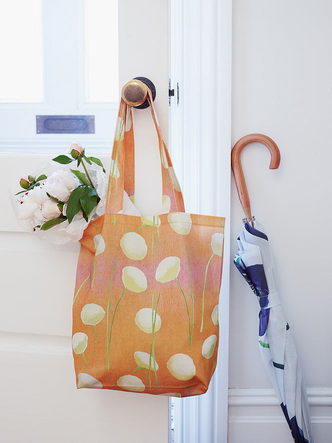 The "Tote Bag: Cream Flowers on Orange," made from stylish cotton canvas and featuring floral patterns, hangs on a doorknob beside a folded umbrella. An oil-painted chrysanthemum bouquet rests against the closed door.