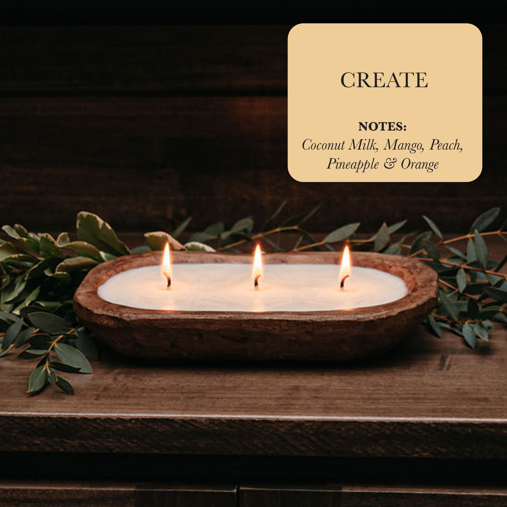 Resting amid vibrant greenery is a luxurious 3-Wick Dough Bowl Soy Candle - Create, featuring three wooden flames. Its label boasts: "Notes - Coconut Milk, Mango, Peach, Pineapple & Orange," with the essence of tropical fruit amplifying its charm.