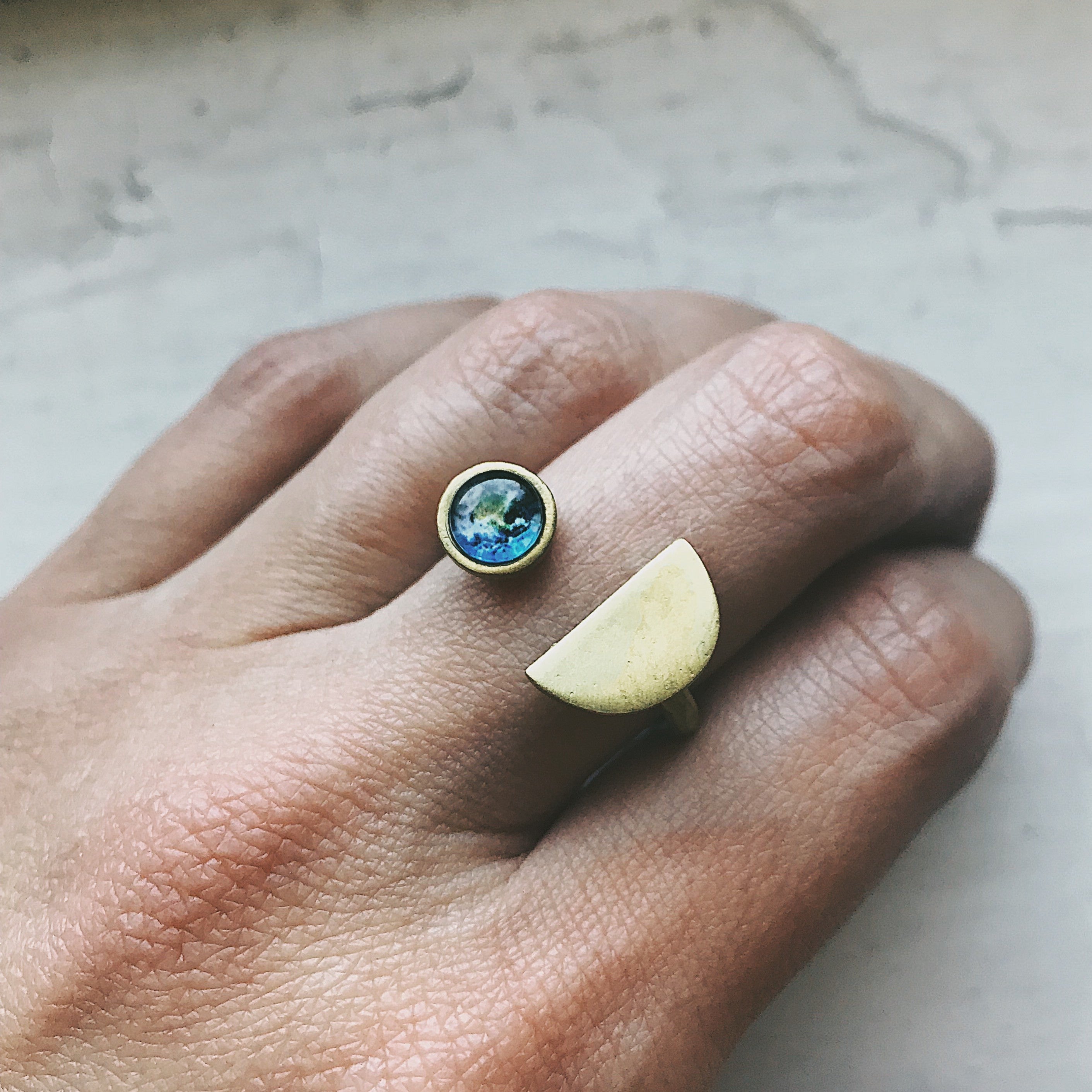 Close-up of a hand adorned with two handcrafted rings: one, the Earth Rise Ring, featuring a small Earth design; and the other showcasing a semi-circular gold shape, capturing the essence of space exploration.