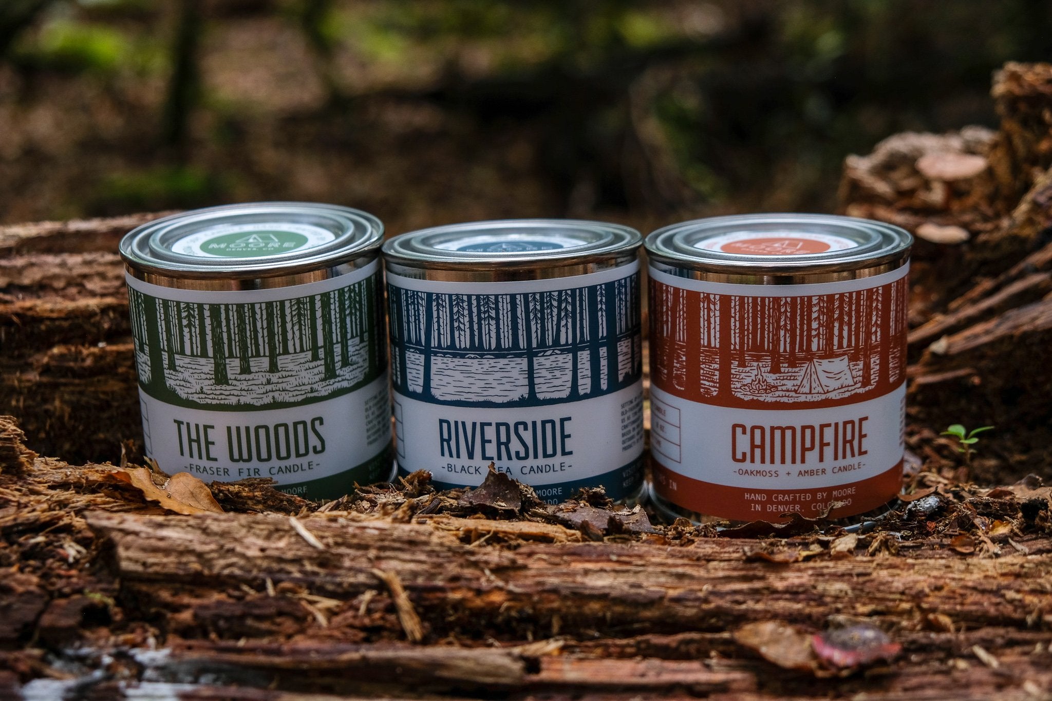 A tin candle called "Campfire Candle" sits on a log, featuring a forest illustration on its label. Handcrafted with natural soy by Moore in Denver, Colorado, this candle releases an inviting outdoor aroma. Lush greenery in the background adds to its rustic charm.