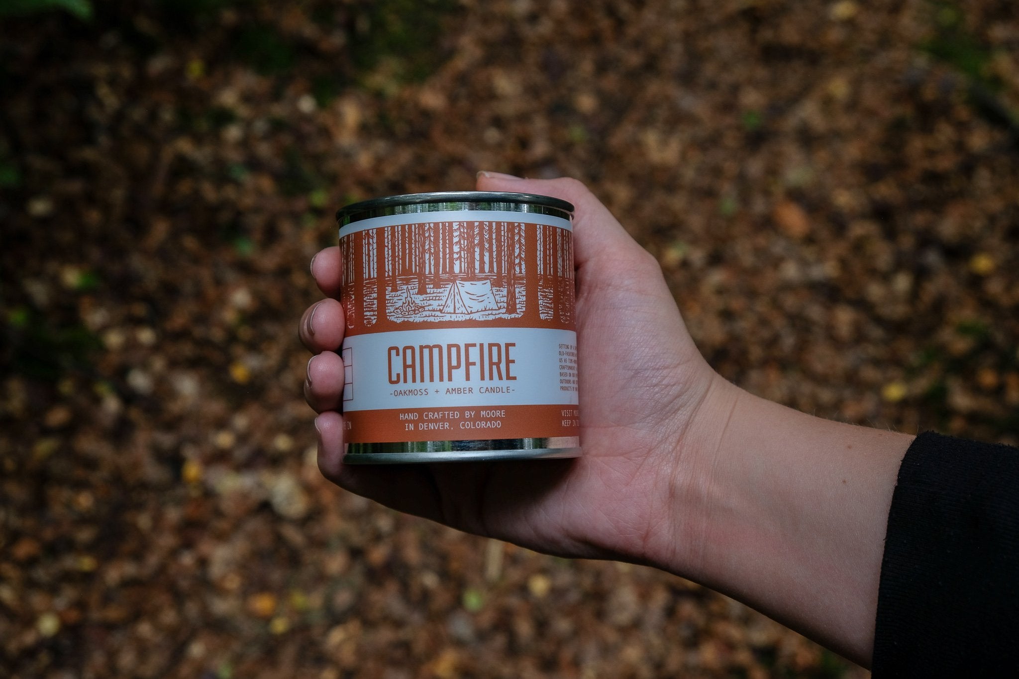 A tin candle called "Campfire Candle" sits on a log, featuring a forest illustration on its label. Handcrafted with natural soy by Moore in Denver, Colorado, this candle releases an inviting outdoor aroma. Lush greenery in the background adds to its rustic charm.