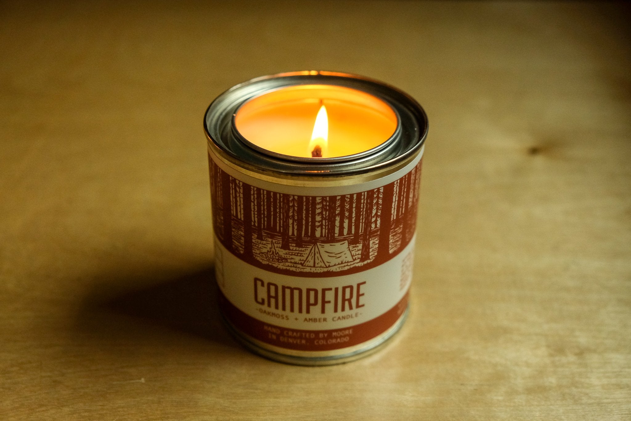 A tin candle called "Campfire Candle" sits on a log, featuring a forest illustration on its label. Handcrafted with natural soy by Moore in Denver, Colorado, this candle releases an inviting outdoor aroma. Lush greenery in the background adds to its rustic charm.