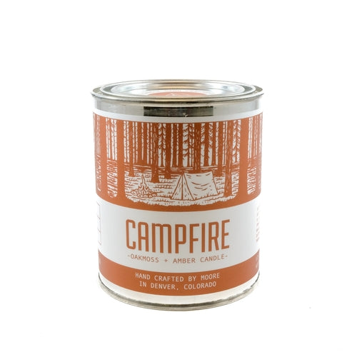 A tin candle called "Campfire Candle" sits on a log, featuring a forest illustration on its label. Handcrafted with natural soy by Moore in Denver, Colorado, this candle releases an inviting outdoor aroma. Lush greenery in the background adds to its rustic charm.