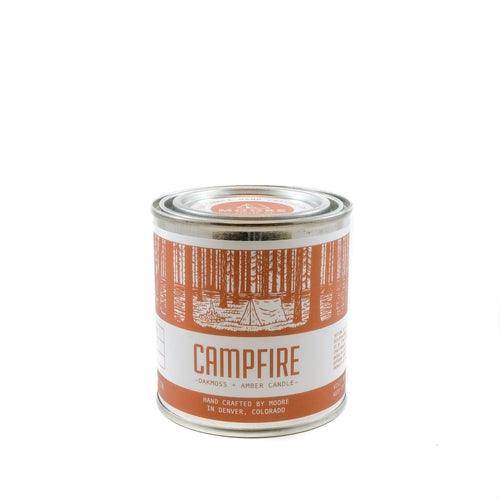 A tin candle called "Campfire Candle" sits on a log, featuring a forest illustration on its label. Handcrafted with natural soy by Moore in Denver, Colorado, this candle releases an inviting outdoor aroma. Lush greenery in the background adds to its rustic charm.