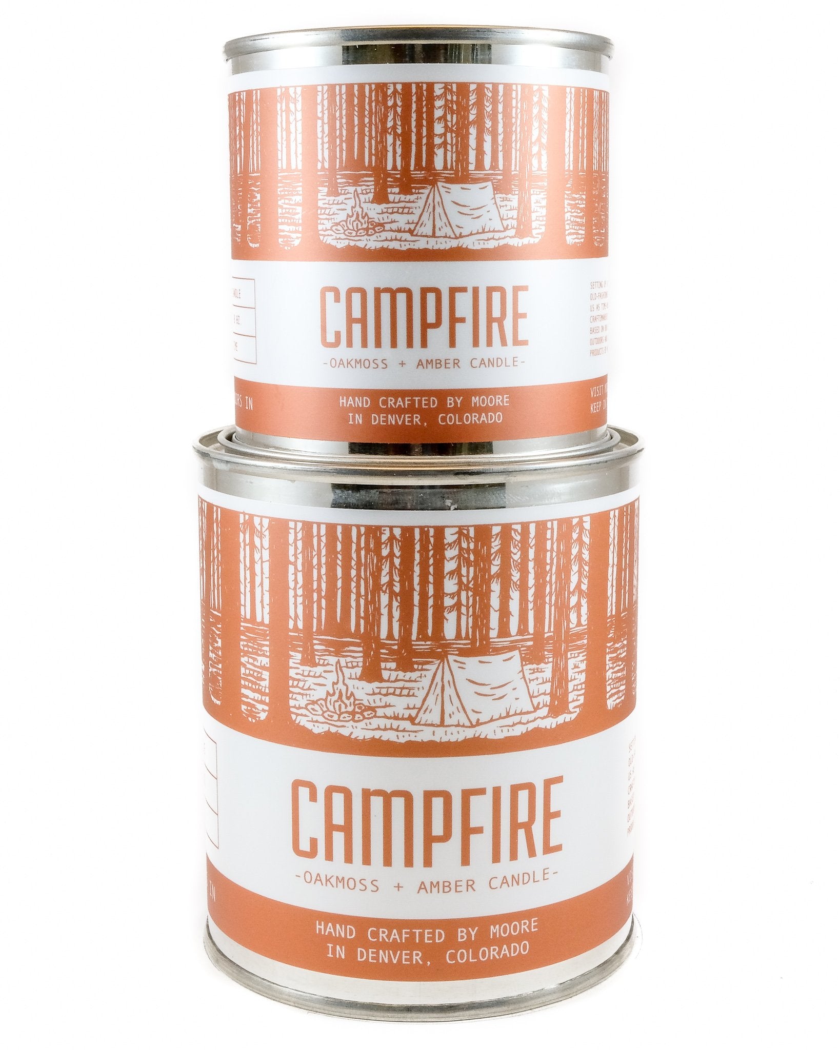 A tin candle called "Campfire Candle" sits on a log, featuring a forest illustration on its label. Handcrafted with natural soy by Moore in Denver, Colorado, this candle releases an inviting outdoor aroma. Lush greenery in the background adds to its rustic charm.