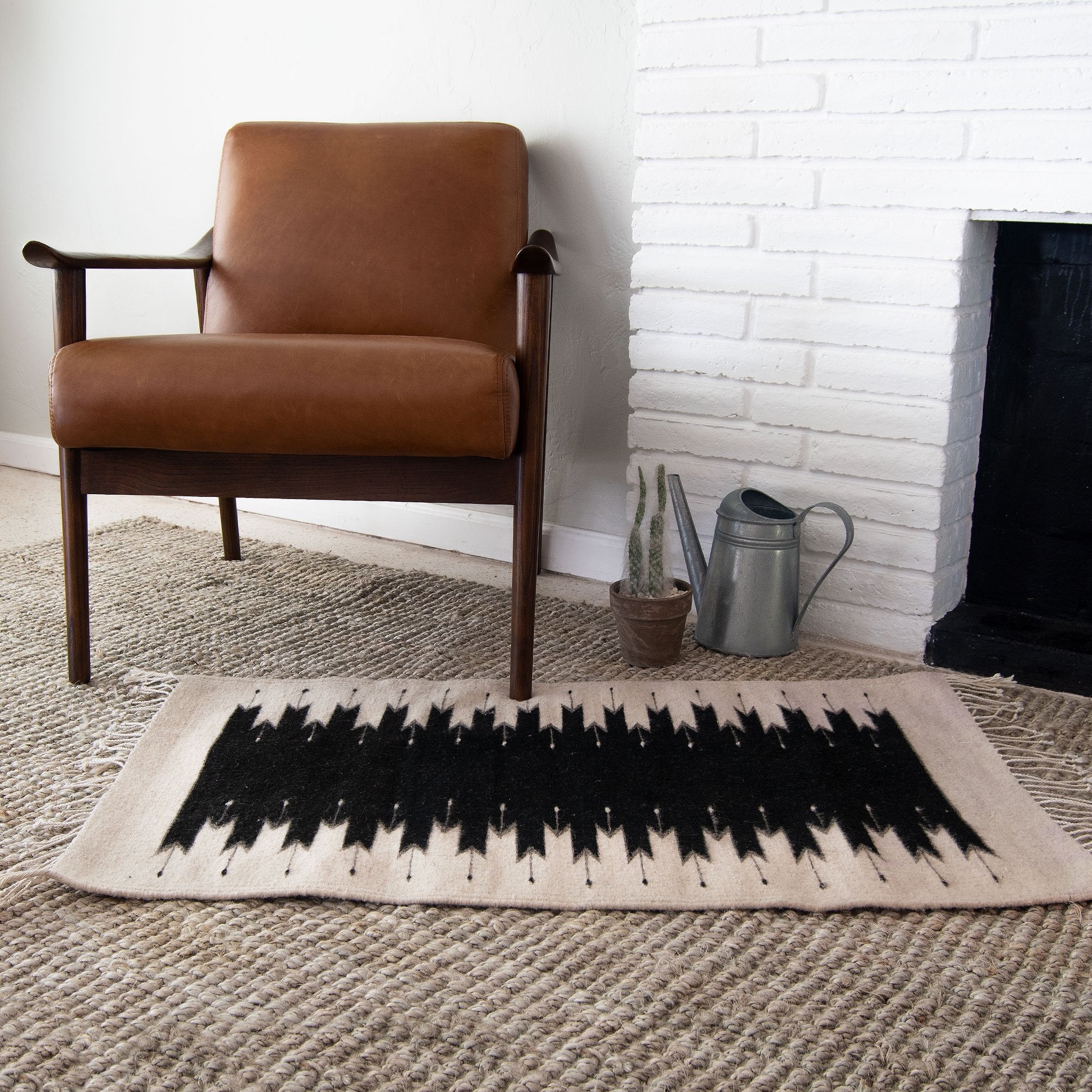 A mid-century modern leather armchair beside a white brick fireplace, with a Beleroó Handmade Southwestern Style Rug. Lightling Pattern in front and a metal watering can on the side.