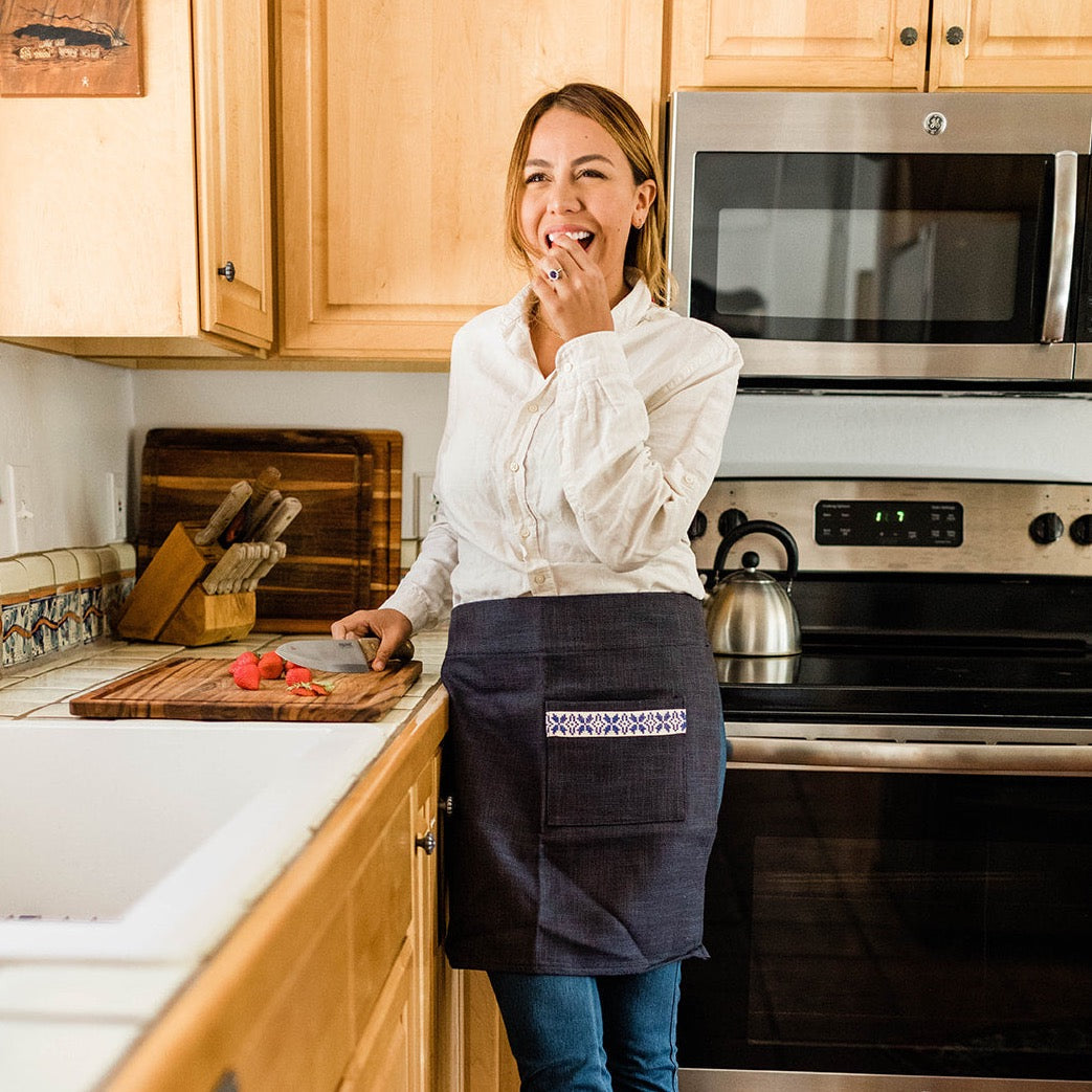 A person stands in a kitchen, hands on hips, wearing the Farah Half Apron - Blue over a white shirt and dark skirt with a blue patterned pocket.